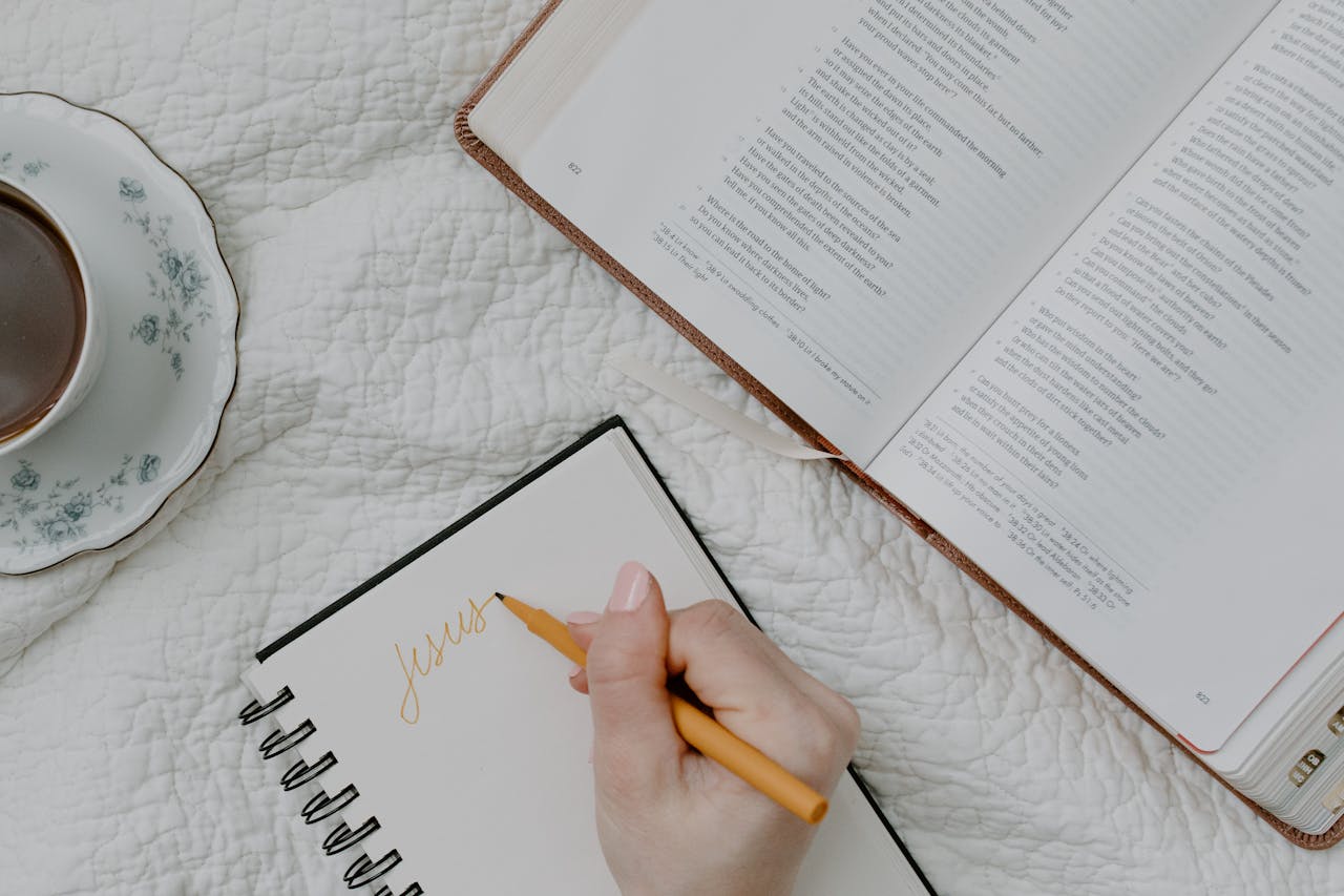 A hand writing in a notebook next to an open Bible and a cup of coffee on a quilted blanket.